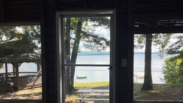 A room with a large window overlooking a lake, a wooden deck, and a picnic table. - Rustic View Cabin sits on a 2.5 acres on beautiful Hubbard Lake.   (Hubbard Lake)