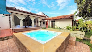 Une piscine avec une terrasse carrelée, un espace extérieur aménagé et couvert avec des rideaux, et une maison avec un toit en tuiles et un jardin.