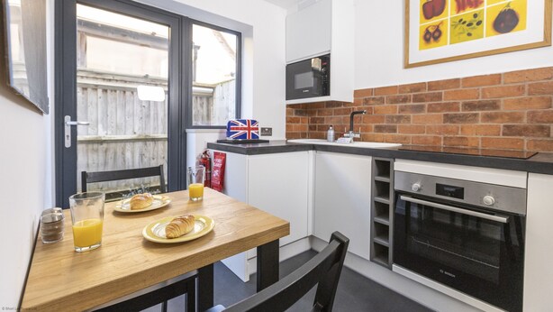 Una cocina compacta con microondas, horno y un lavamanos pequeño. Hay una mesa de desayuno con croissants y jugo, y una decoración con la bandera británica.