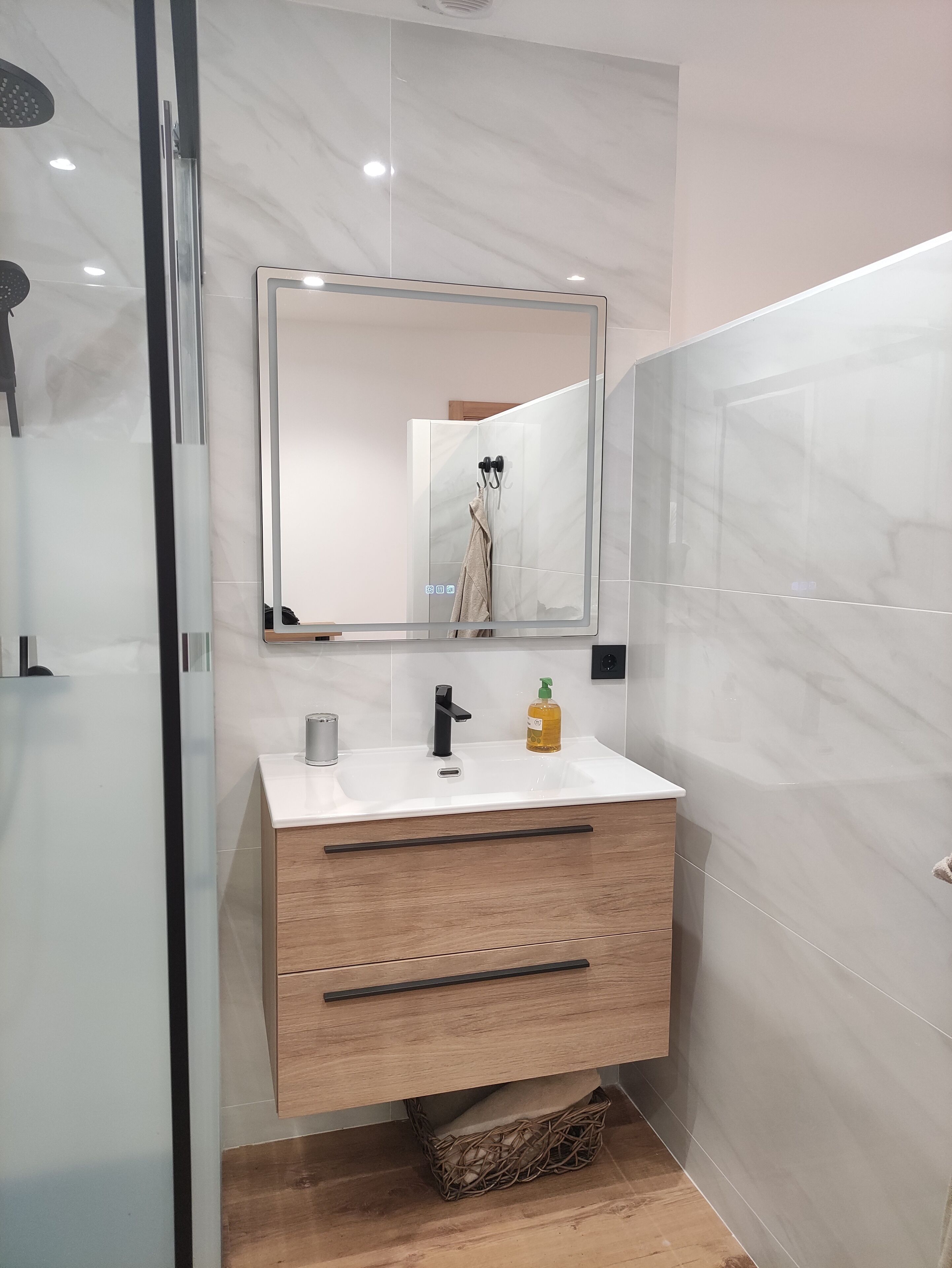 Une salle de bain moderne avec une douche à parois de verre, un lavabo blanc doté d’un robinet noir et un meuble-lavabo en bois avec des tiroirs.