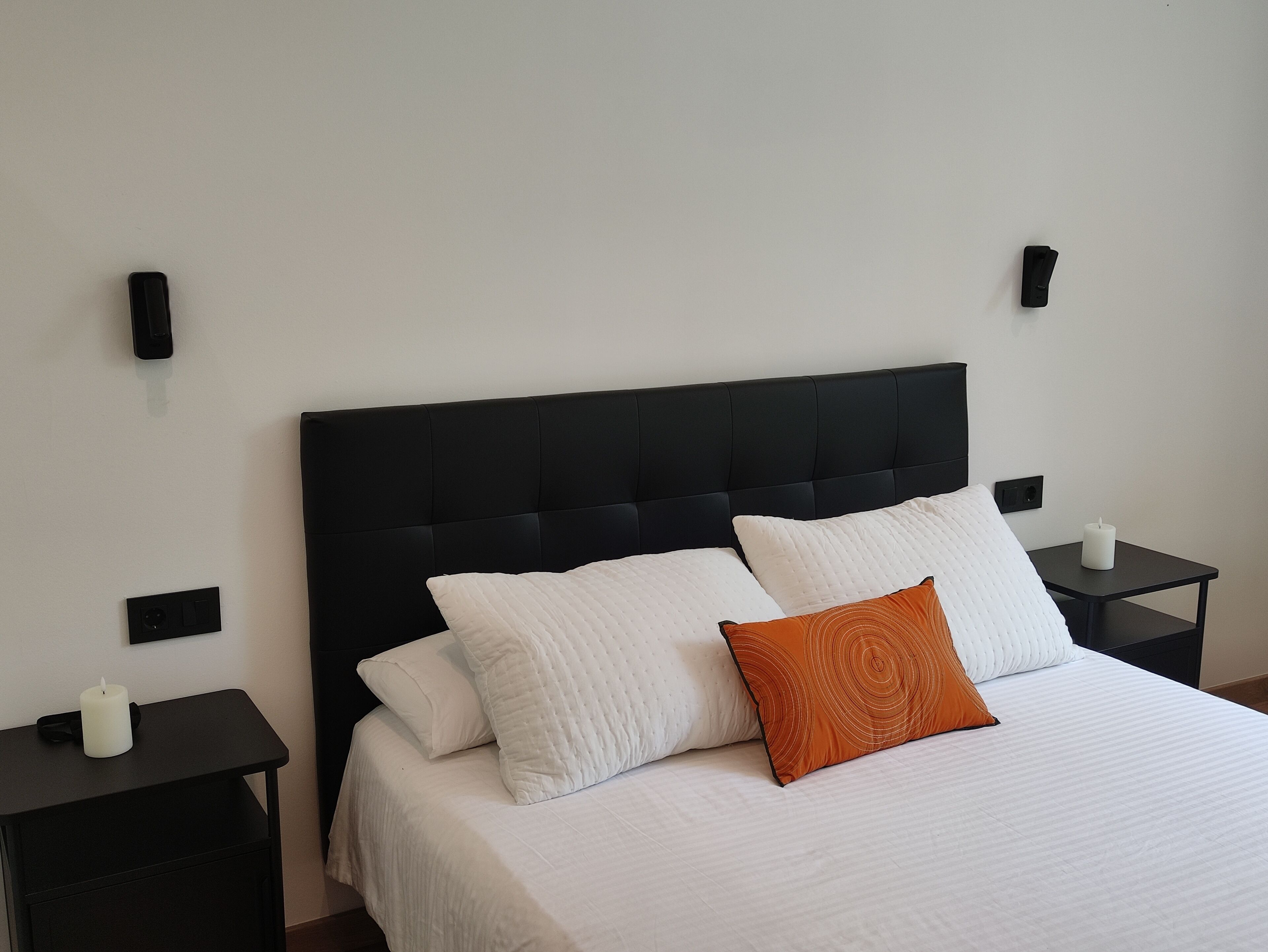 Een net opgemaakt bed met witte en oranje kussens, een zwart hoofdbord en twee nachtkastjes met kaarsen.