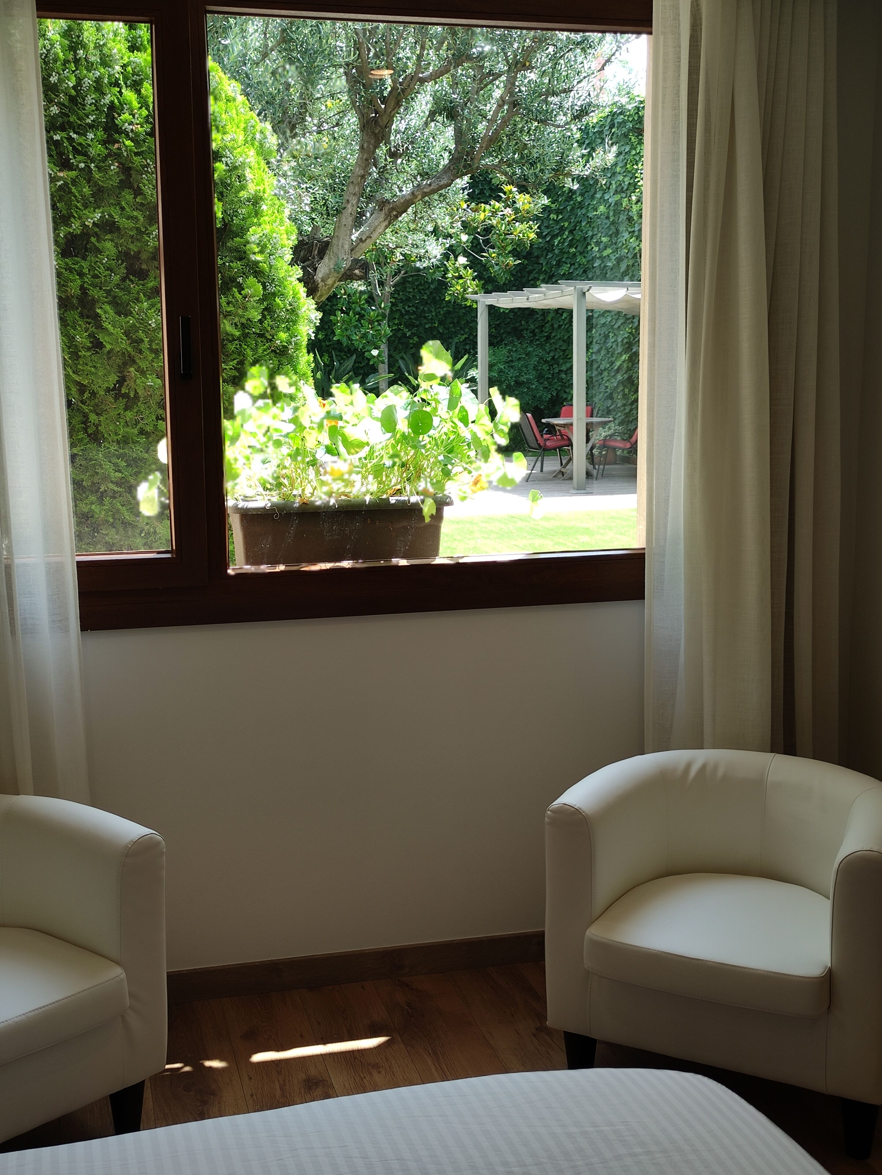 Une pièce dotée d’une grande fenêtre donnant sur un jardin, de deux fauteuils blancs, d’un lit et d’un plancher en bois.