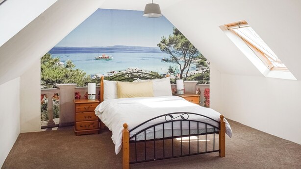 Ein Schlafzimmer mit Bett, Nachttisch, Lampe und Meerblick durch ein Fenster.