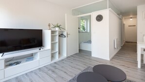 Una sala de estar moderna con un televisor de pantalla plana, estanterías blancas, una pequeña zona de estar con dos mesas redondas y una escalera que lleva a otra habitación.