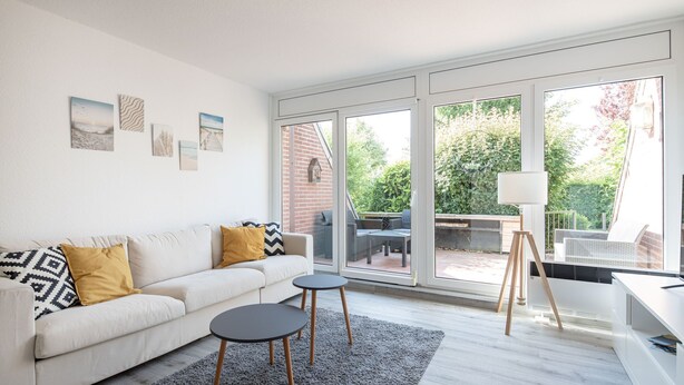 Una sala de estar moderna con un sofá blanco, dos mesitas, un tapete gris y un amplio ventanal con vistas a un jardín y un muro de ladrillo.