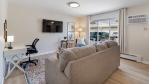 Een moderne woonkamer met een grote beige bank, een bureau met stoel, een flatscreen televisie en een groot raam met gordijnen.