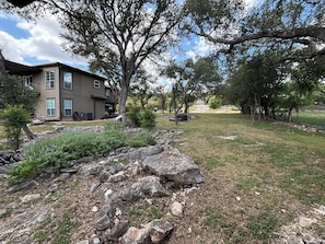 A residential area with a house, a stone retaining wall, and a grassy yard with trees. - Lake Forest Escape: Secluded Retreat with Breathtaking Lake & Country Views (Canyon Lake)
