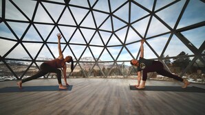 Twee personen beoefenen yoga in een ruimte met een geometrisch glazen wand en een houten vloer.