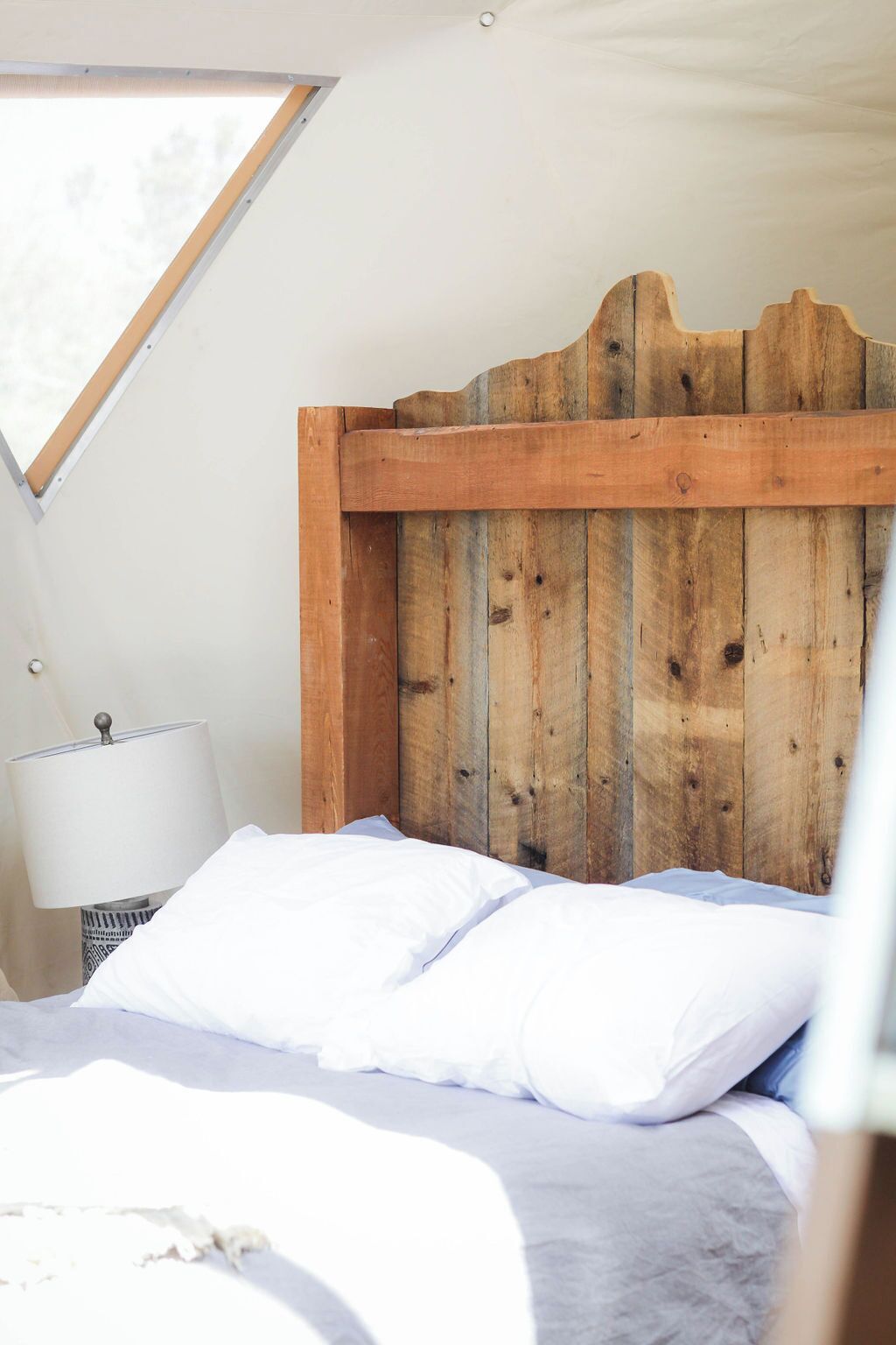 A bedroom with a wooden headboard, a bed with white bedding, a bedside lamp, and a window with a view of trees.