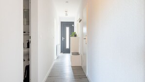 Ein schmaler Flur mit Holzdielen, weißen Wänden und einer Tür am Ende, die in einen Raum führt.