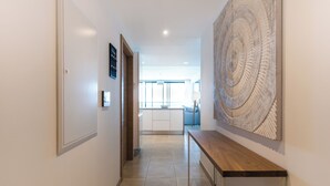 Un couloir avec un banc en bois, une grande œuvre d’art murale tissée et une vue sur un espace cuisine moderne.