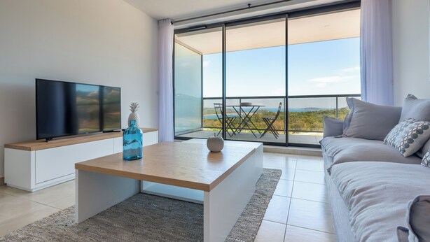 Un salon moderne doté d’un téléviseur à écran plat, d’une table basse en bois, d’un canapé et d’une grande porte-fenêtre coulissante donnant sur un balcon avec vue sur le paysage.