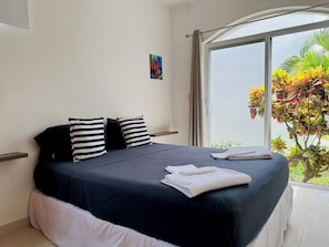 A neatly made bed with a dark blue bedspread and striped pillows, a small painting on the wall, and a view of a tree through a large window.
