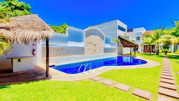 Una piscina con una pared de piedra, una estructura de techo de paja y un camino pavimentado.