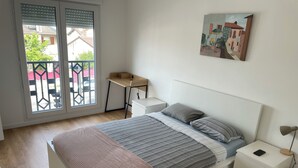 A bedroom with a bed, bedside tables, a desk, and a window with a view of a red fence and buildings. - Appartement Cosy : Paris/cdg/ Parking Prive (Le Blanc-Mesnil)