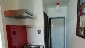 A compact kitchen with a stove, sink, and a red backsplash. A hallway leads to a door at the end. - Studio VUE MER (Agde)