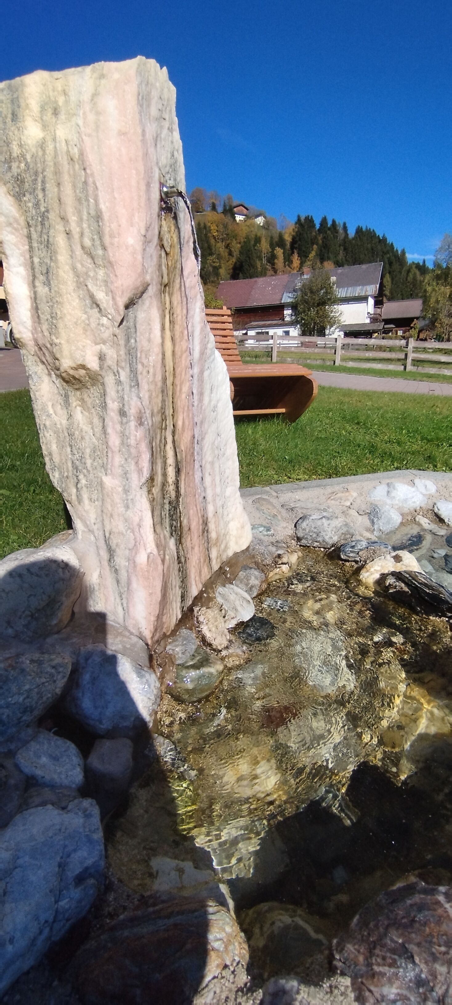 Una estructura de piedra con un elemento acuático, rodeada de césped y casas al fondo.
