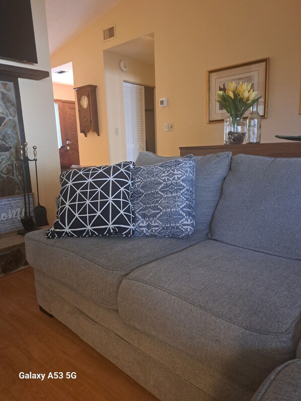 A living room with a grey sofa, patterned cushions, a clock, a vase with flowers, and a framed picture on the wall.