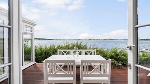 En udendørs terrasse med et hvidt bord og stole, med udsigt over en sø med både og grønt i baggrunden.