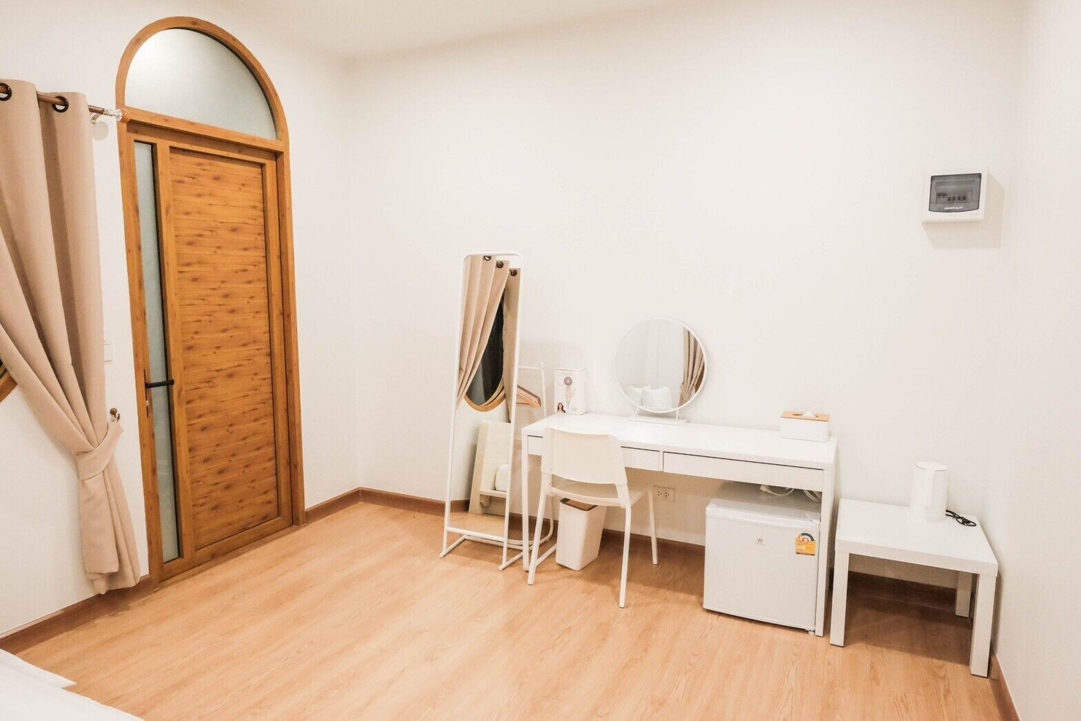A small, tidy room with a wooden door, a white desk with a chair, a small cabinet, and a mirror.