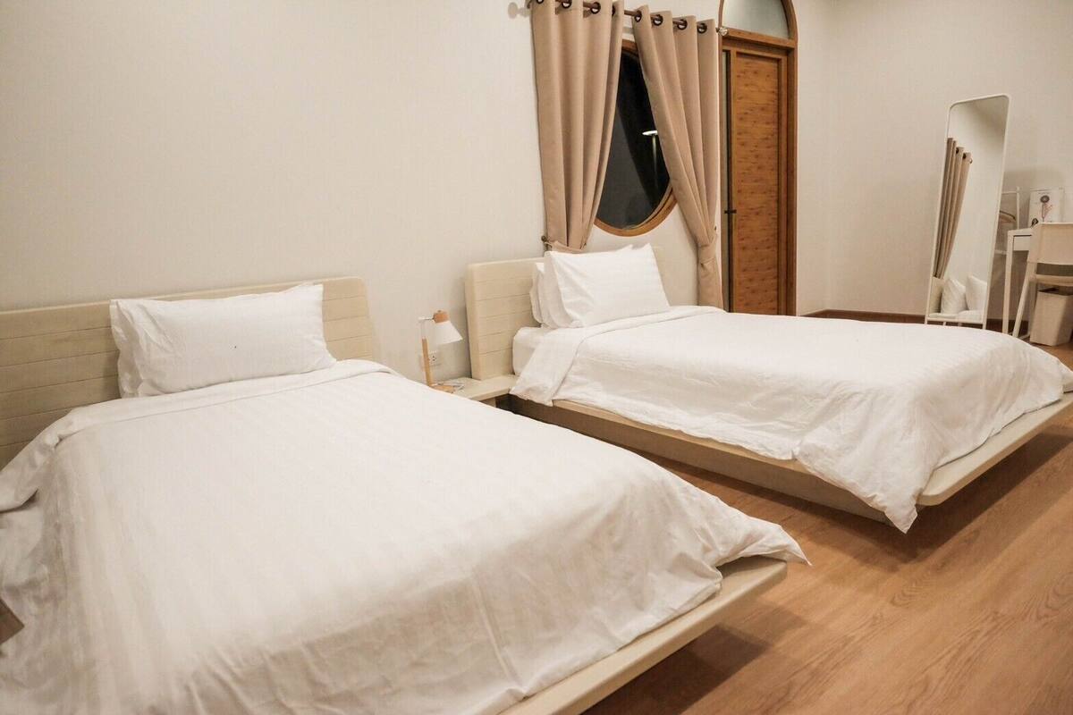 A hotel room with two single beds, a wooden floor, a window with curtains, and a mirror.