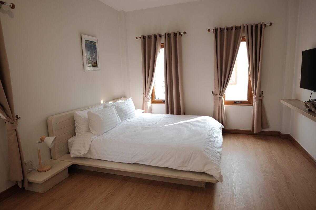 A neatly arranged bedroom with a bed, curtains, a bedside lamp, a wall-mounted TV, and a framed picture.