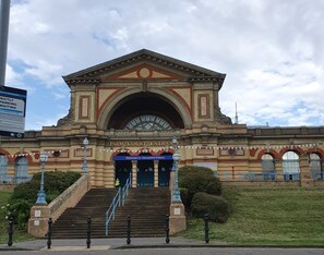 A grand entrance with steps leading up to a large archway, a sign welcoming visitors to Alexandra Palace, and a clear sky above.
