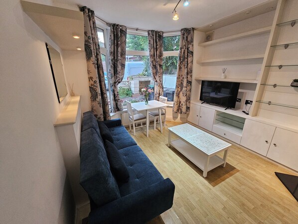 A modern living room with a dark blue sofa, a white coffee table, and a TV on a white cabinet. There is a dining area with a table and chairs, and large windows with floral curtains.