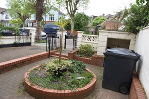 A residential garden with a brick border, a black trash bin, and a white wall.