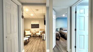 A hallway with wooden flooring leading to a bedroom with a bed and a nightstand. There is a seating area with chairs and a table, and a decorative wall piece above the seating area. - Beachy Getaway Near Newport & Beaches (Middletown)