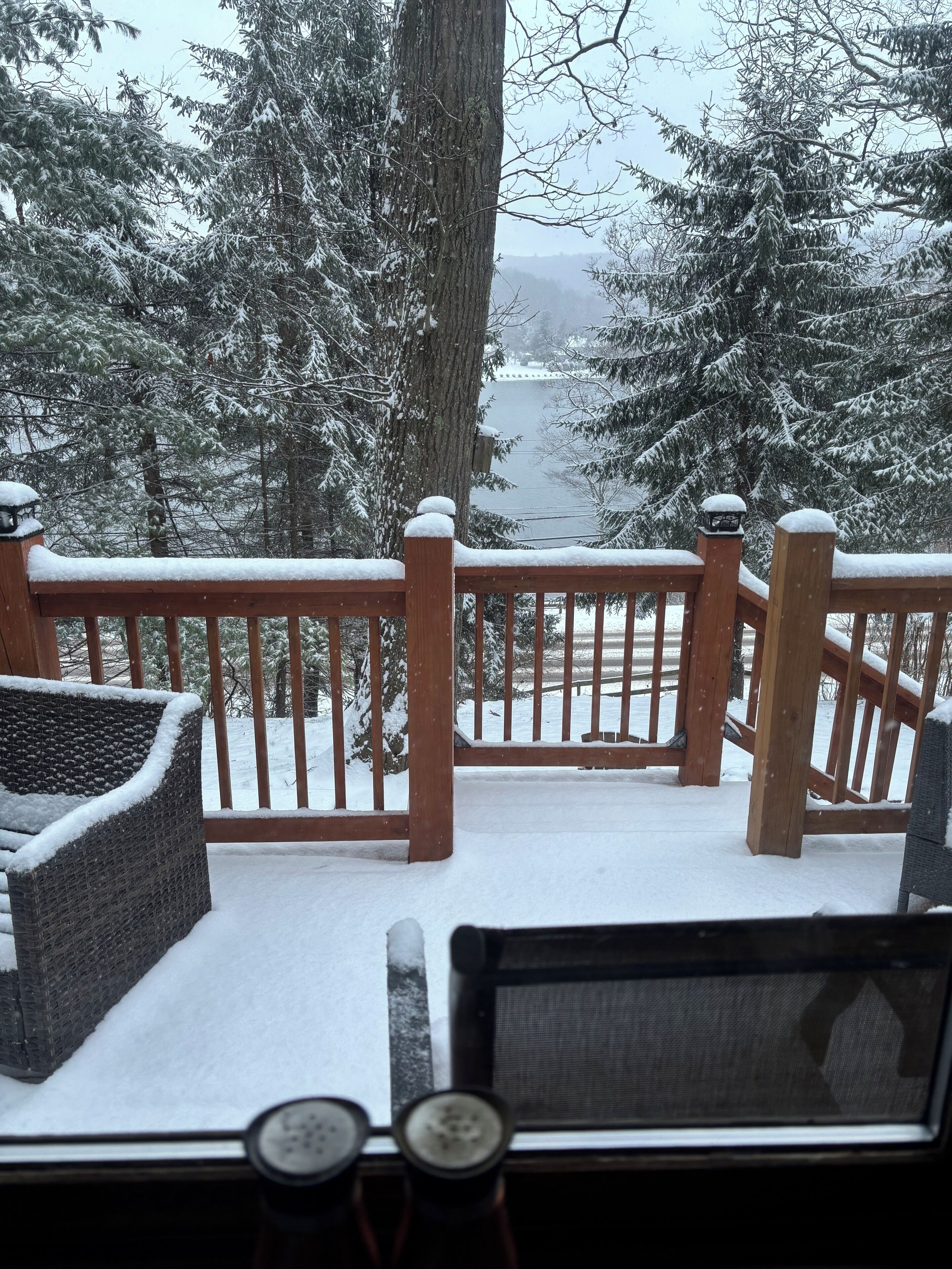 A deck with a wooden railing and wicker furniture, overlooking a snowy landscape with trees and a body of water.