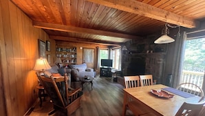 Indretning i en hyggelig hytte med træloft, pejs, sofa, fjernsyn og spisebord med stole.