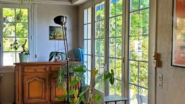 A room with a large window, a hanging light fixture, a wooden cabinet, and a black sofa.