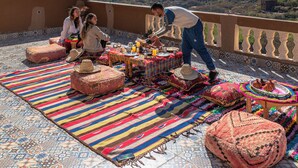 Una terrassa amb una taula preparada per a dinar, envoltada de catifes i otomans de colors, amb vista a un paisatge de muntanyes.