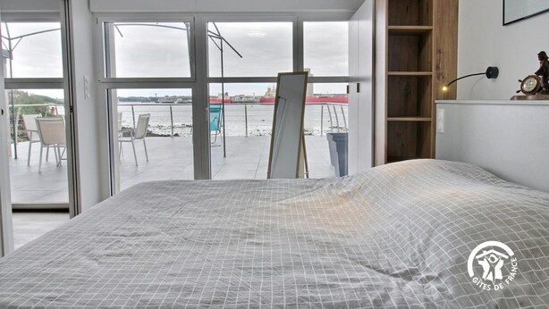 Une chambre avec un grand lit, une étagère en bois et une vue sur un balcon agrémenté de chaises et d’une table.