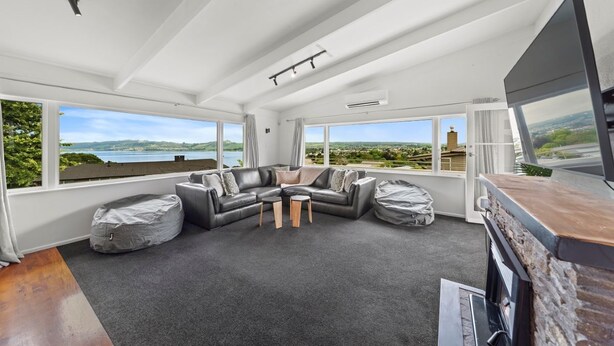 A spacious living room with a large sectional sofa, a coffee table, and a TV. The room has large windows offering a view of a lake and greenery. - Hilltop House Lake Views (Taupo)