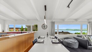 A modern kitchen with a central island, dining area with a white table and chairs, and a view of a lake through large windows. - Hilltop House Lake Views (Taupo)
