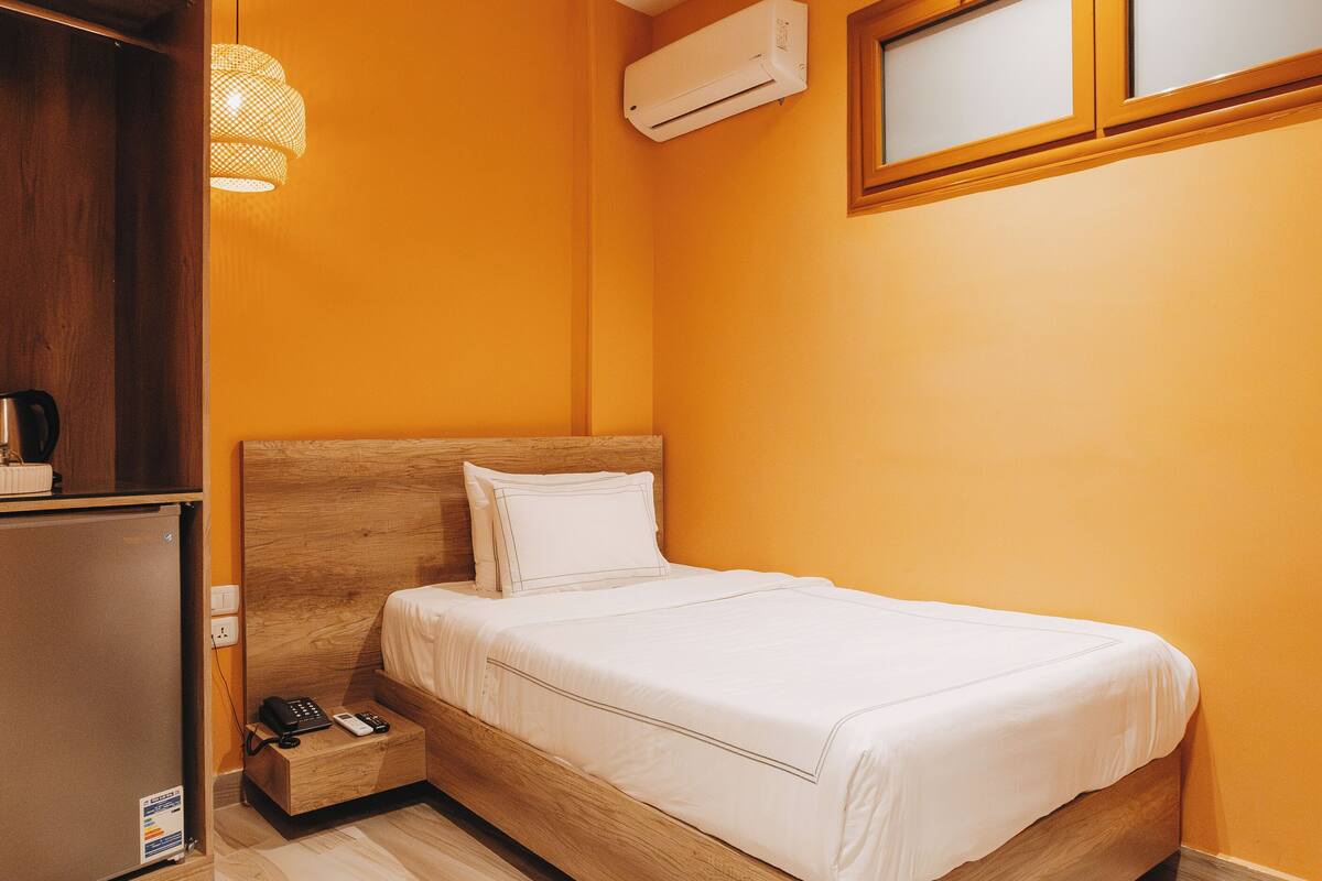 A hotel room with a wooden bed, a small bedside table with a phone and a lamp, an air conditioning unit, and a window with blinds.