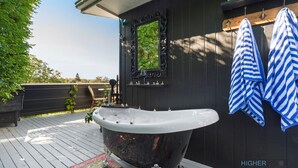 Een zwart bad op een houten terras, met een donkere houten wand, een spiegel en twee gestreepte handdoeken aan een rek.