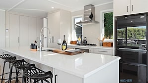 Een moderne keuken met witte kasten, een groot eiland met barkrukken, een roestvrijstalen afzuigkap en uitzicht op groen buiten.