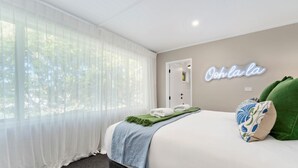 Een hotelkamer met een bed, een neonbord aan de muur, een raam met dunne gordijnen en een groen kussen.