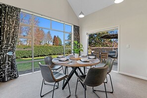 Uma área de refeições com uma mesa redonda posta para seis pessoas, rodeada por cadeiras pretas com detalhes dourados. Grandes janelas oferecem vista para um jardim e árvores.