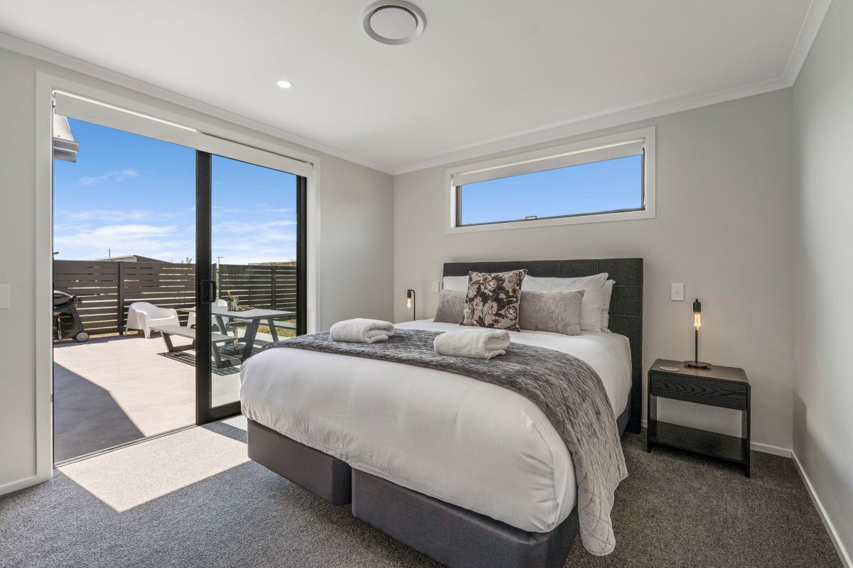 A modern bedroom with a large bed, a small bedside table, a window, and a sliding glass door leading to an outdoor area.