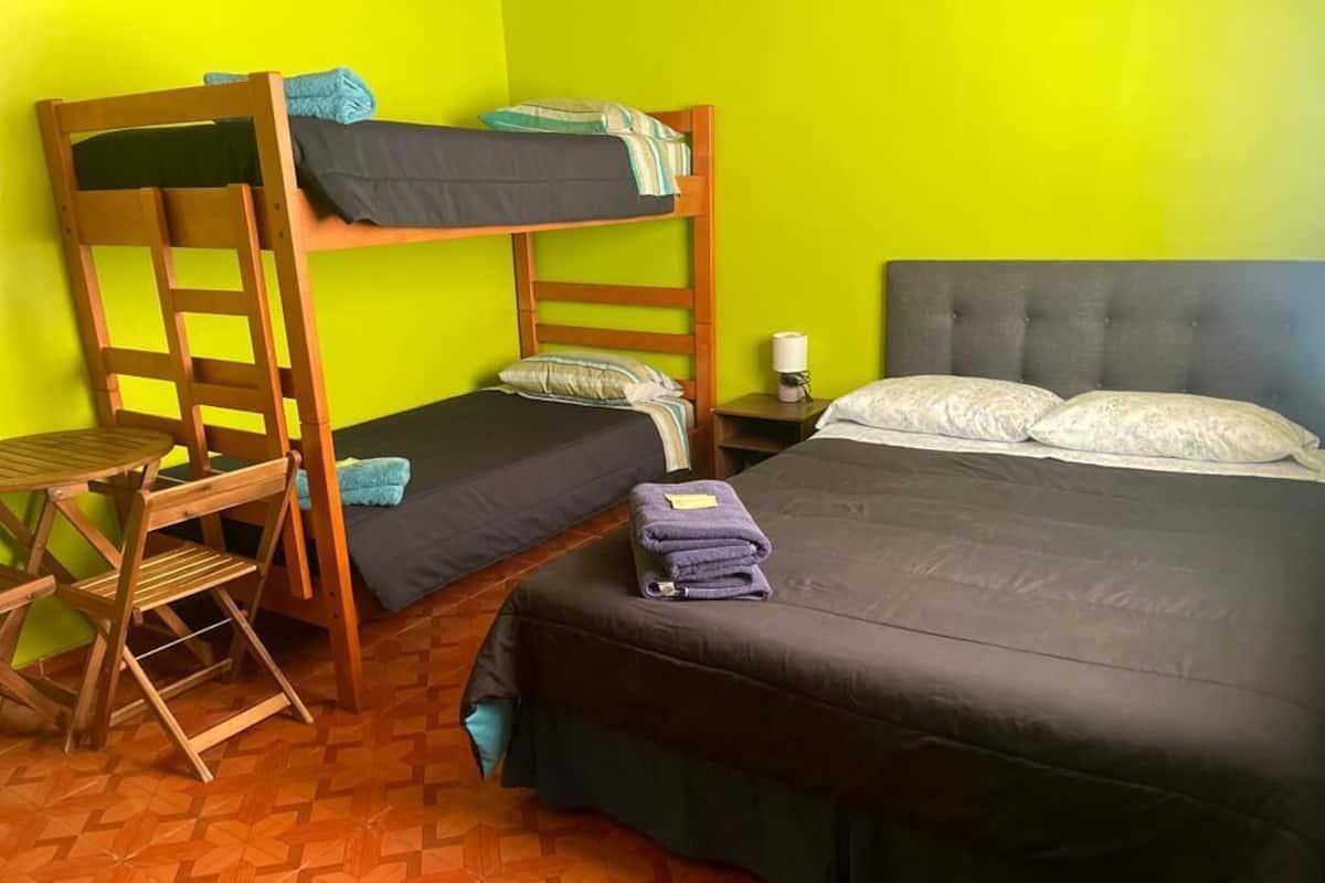 Une chambre avec un lit superposé, un lit simple, une chaise en bois, une petite table et une table de chevet avec une lampe.