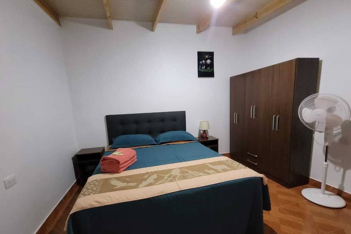 Une chambre à coucher comprenant un lit, une table de chevet, une armoire et un ventilateur sur pied.