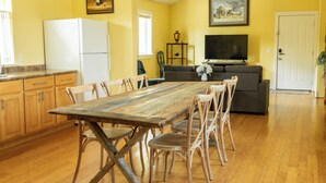 Ein Essbereich mit Holztisch und Stühlen, eine Küche mit Kühlschrank sowie ein Wohnzimmer mit Sofa und Fernseher.