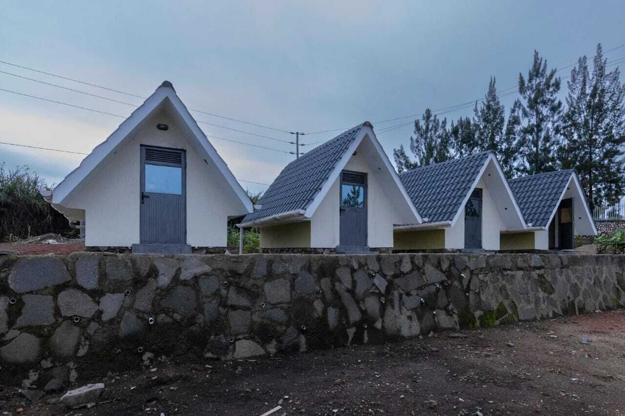 A row of three small, white, triangular buildings with grey roofs, each with a door and a window, set against a stone wall and surrounded by trees.
