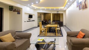 A modern living room with a glass coffee table, brown sofas, a dining area with a wooden table and chairs, and a TV mounted on the wall. - Kaije Country Cottages (Fort Portal)