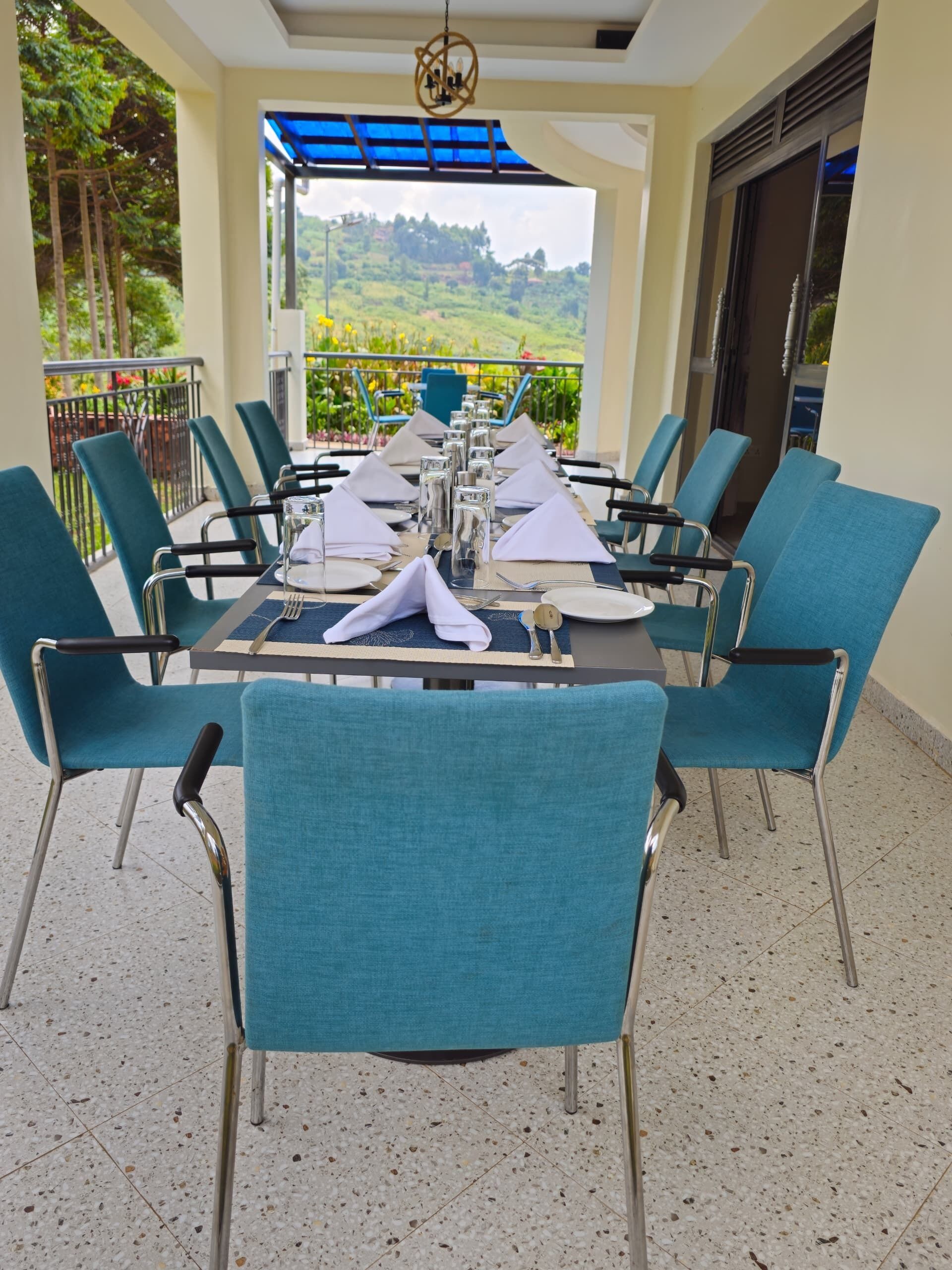 A dining area with a table set for six, surrounded by teal chairs, with a view of greenery and hills in the background.
