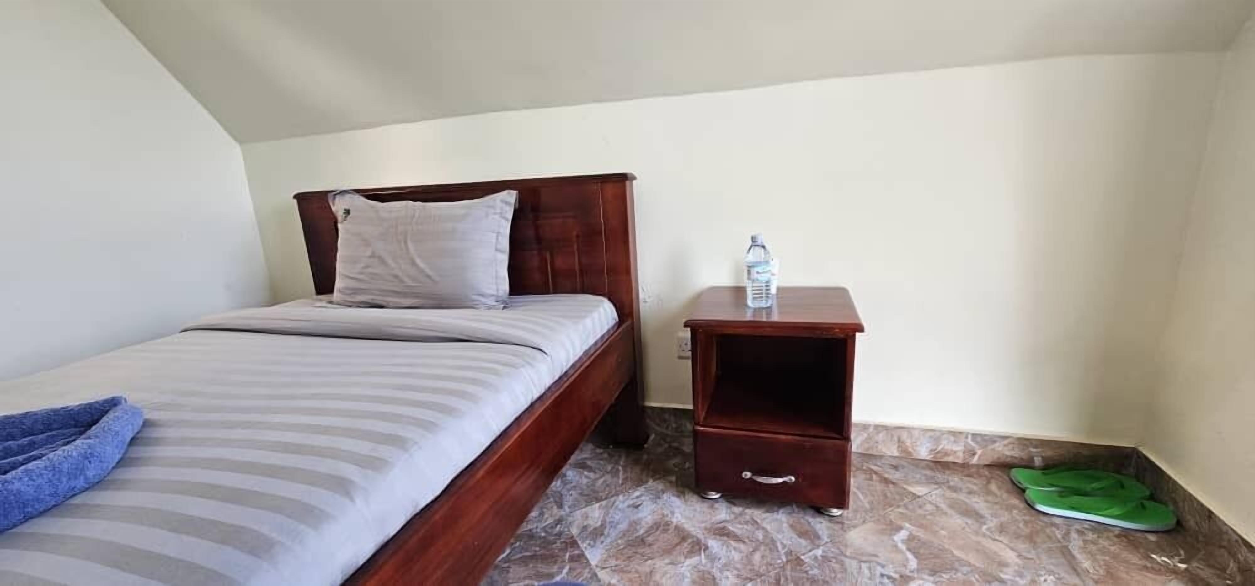 A single bed with a wooden headboard and a nightstand with a water bottle and slippers.